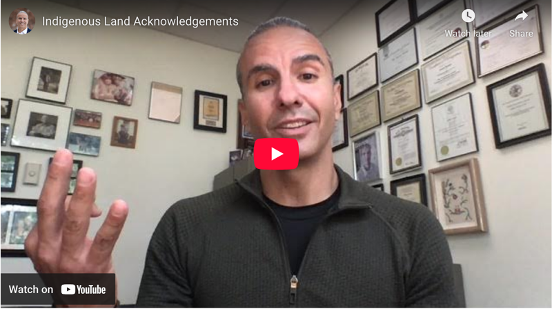 The image shows a man in a room with framed documents on the wall. He is wearing a dark green sweater and is gesturing with his hands. The video is titled "Indigenous Land Acknowledgements". The video is on YouTube.